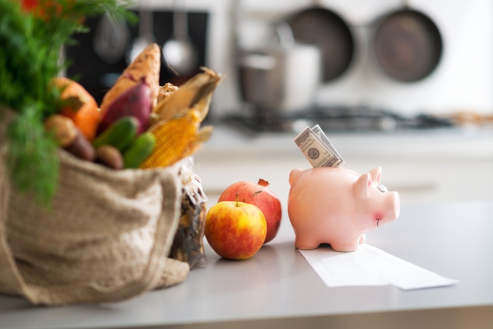 Piggy bank with money in the top on a table next to a bag of fruit and vegetables