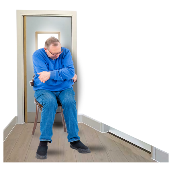 An older man in a blue sweatshirt and jeans sitting on a chair in a narrow hallway.