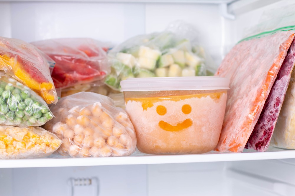 Portions of frozen vegetables and soup in a freezer, with smiley face drawn in frost on a tub