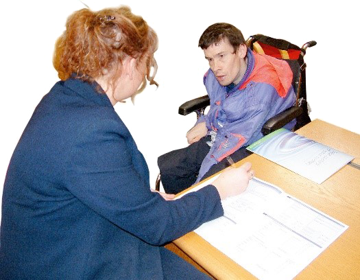 A person is sitting in a wheelchair. Next to him is a person filling in forms at an office desk.