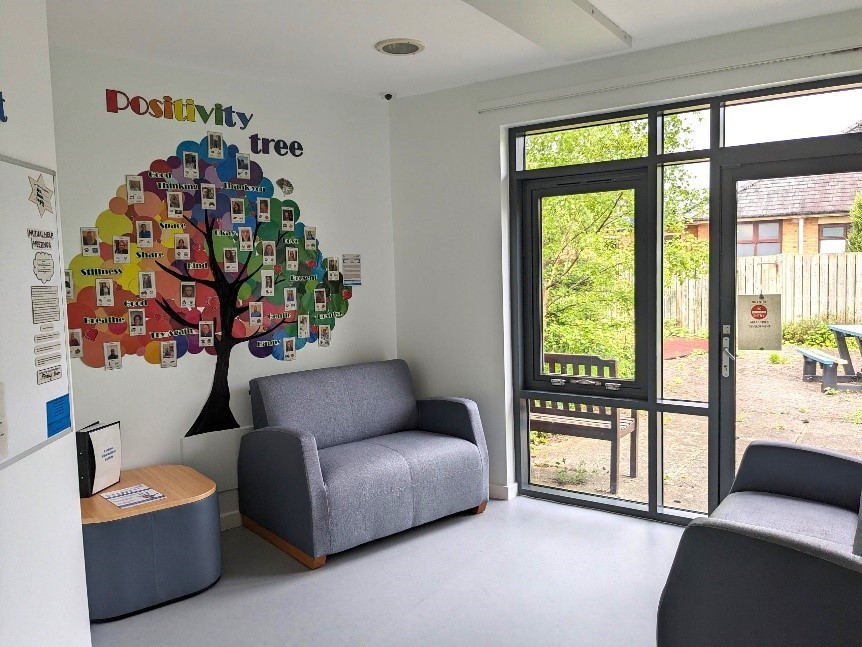 A room with 2 sofas, coffee table and patio doors. There is a brightly coloured tree decorating one wall.