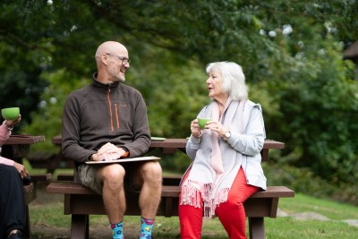 A man and older woman sitting on a park bench chatting, the woman is holding a cup of tea